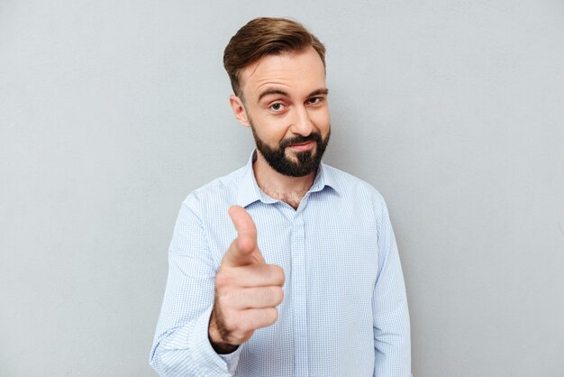 Uomo barbuto sorridente sleale in vestiti di affari che indica alla macchina fotografica