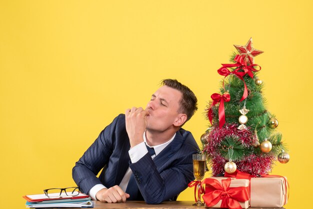 Uomo beato vista frontale che fa gesto di bacio cheff seduto al tavolo vicino albero di Natale e regali su sfondo giallo