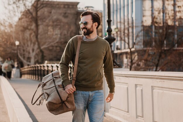 Uomo bello alla moda hipster che cammina nella strada della città con borsa in pelle che indossa tuta e occhiali da sole, tendenza stile urbano, giornata di sole, viaggiatore felice sorridente