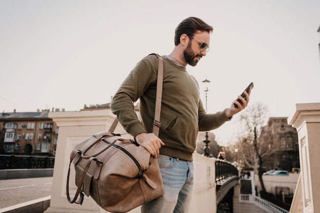 Uomo bello alla moda hipster che cammina nella strada della città con borsa in pelle utilizzando l'applicazione di navigazione del telefono, viaggio con indosso felpa e occhiali da sole, tendenza in stile urbano