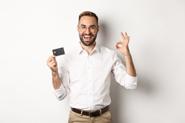 Uomo bello che mostra la sua carta di credito e segno giusto, raccomandando banca, in piedi Copia spazio