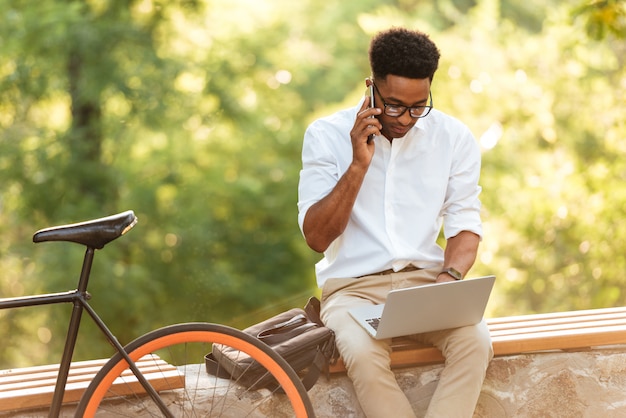 Uomo bello che per mezzo del computer portatile che parla dal telefono.