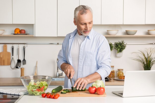 Uomo bello che sta alla cucina facendo uso del computer portatile e della cottura