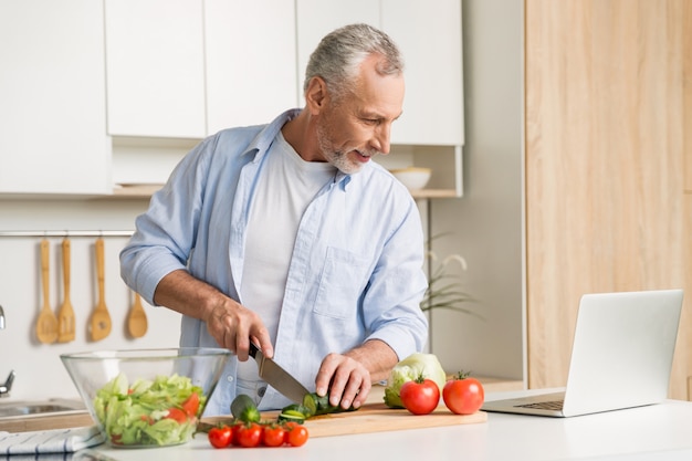 Uomo bello che sta alla cucina facendo uso del computer portatile e della cottura