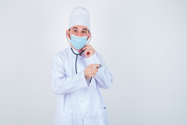 Uomo bello che tiene i pezzi dell'orecchio dello stetoscopio nelle orecchie come se stesse ascoltando in camice medico bianco, maschera e guardando felice, vista frontale.