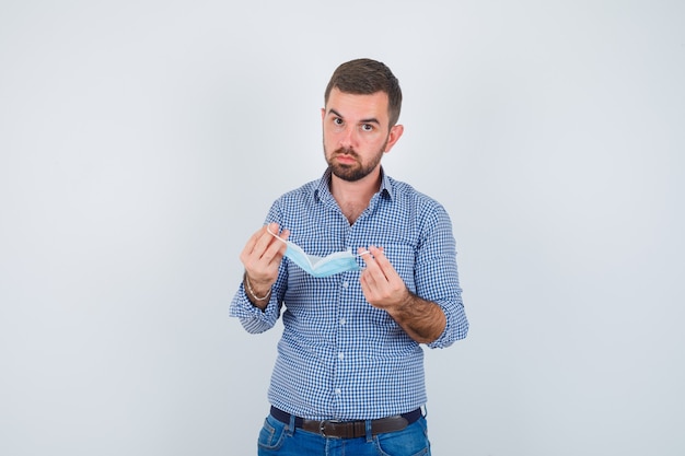 Uomo bello che tiene la maschera in camicia, jeans e che sembra serio. vista frontale.