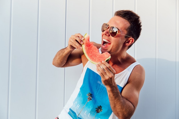 Uomo bello divertente in occhiali da sole mangiare anguria gustoso, ridendo forte