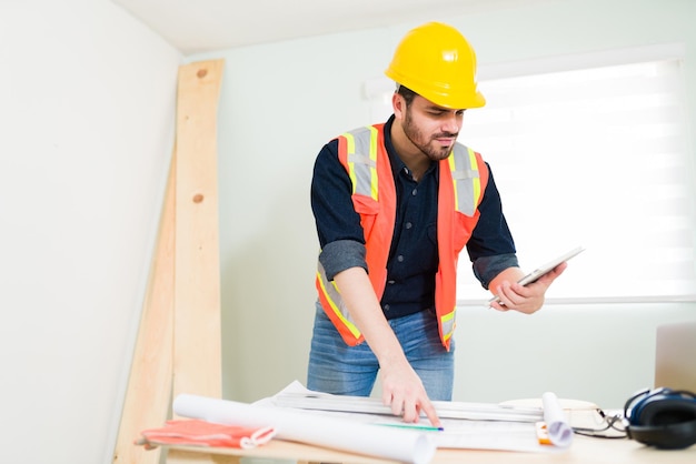 Uomo bello e appaltatore con un casco di sicurezza che controlla i progetti di costruzione mentre guarda un tablet