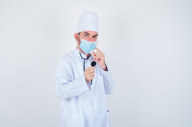 Uomo bello in camice da laboratorio medico bianco, maschera che tiene gli auricolari dello stetoscopio nelle orecchie come se stesse ascoltando e guardando concentrato, vista frontale.