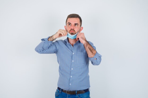 Uomo bello in camicia, jeans, maschera che tiene la maschera sotto il mento e che sembra serio, vista frontale.