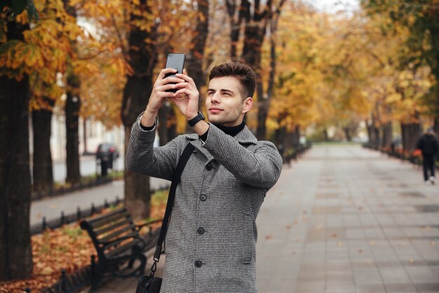 Uomo bello in cappotto che prende foto di bei alberi di autunno facendo uso del suo cellulare moderno mentre camminando nel parco vuoto