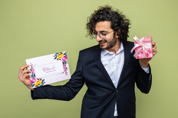 Uomo bello in vestito che tiene la cartolina d'auguri e presente guardando la carta sorridente felice e positivo che celebra allegramente la giornata internazionale della donna 8 marzo in piedi su sfondo verde