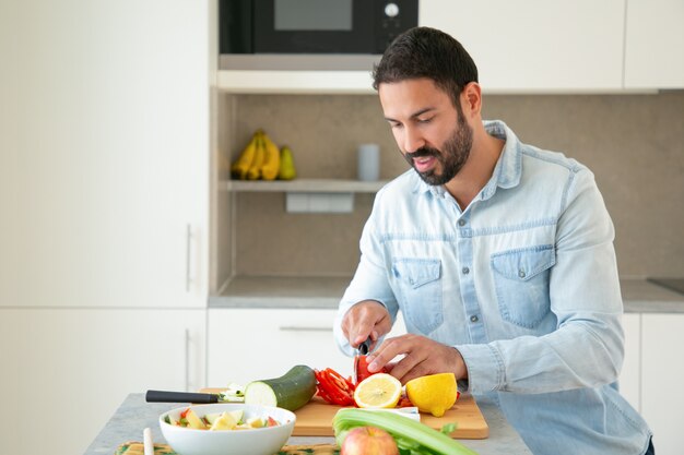 Uomo bello positivo che cucina insalata, tagliare le verdure fresche sul tagliere in cucina. Colpo medio, copia spazio. Concetto di cibo sano