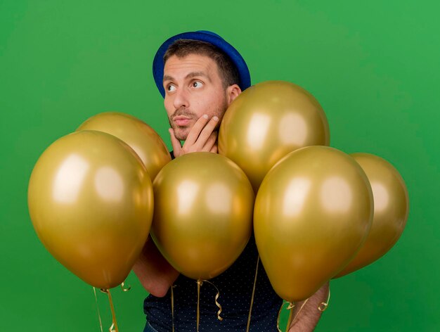 Uomo bello premuroso che indossa il cappello blu del partito mette la mano sul mento sta con gli aerostati dell'elio che esaminano il lato isolato sulla parete verde con lo spazio della copia