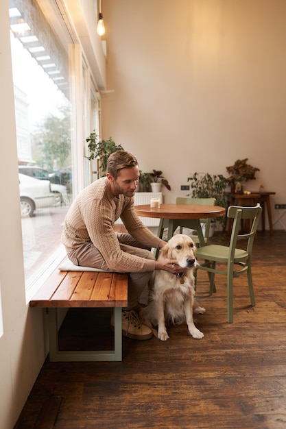 Uomo bello seduto in un caffè con il suo cane un tizio beve caffè e accarezza un golden retriever