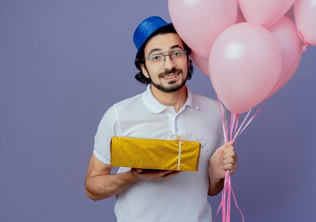 Uomo bello sorridente con gli occhiali e cappello blu che tiene palloncini e confezione regalo isolato su viola