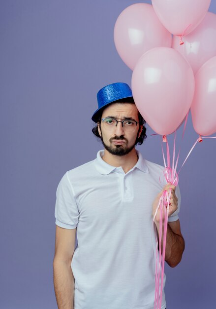 Uomo bello triste con gli occhiali e cappello blu che tiene palloncini isolati sulla porpora