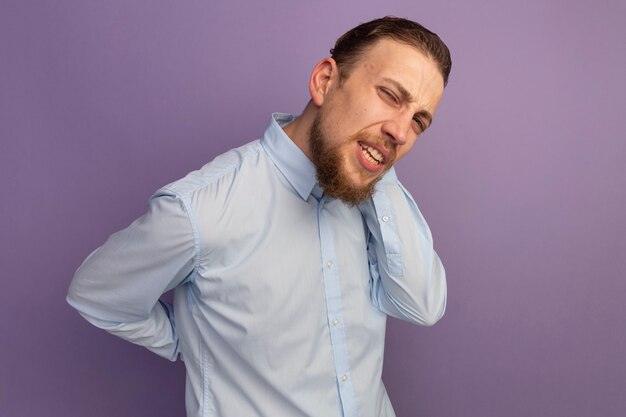 Uomo biondo bello dolorante trattiene la schiena e il collo dietro isolato sulla parete viola