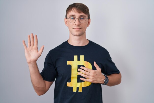 Uomo biondo caucasico che indossa una maglietta bitcoin che giura con la mano sul petto e il palmo aperto, facendo un giuramento di promessa di fedeltà