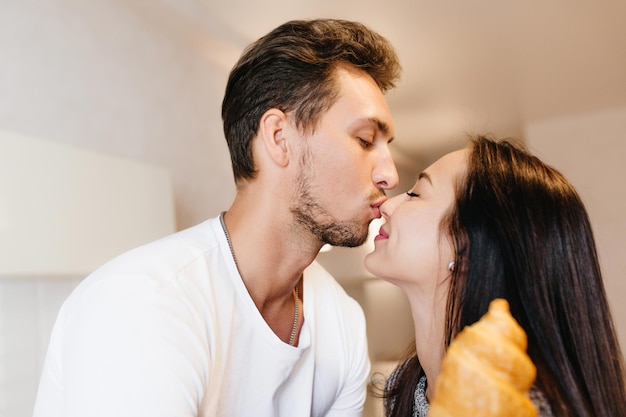 Uomo brunetta barbuto che bacia dolcemente la ragazza nel naso. Ritratto al coperto di una magnifica ragazza dai capelli neri che tiene in mano un croissant che si diverte con il marito nel fine settimana.