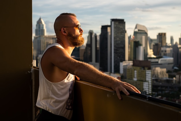 Uomo brutale barbuto bello caucasico che gode del tramonto e vista sulla città di Bangkok dal piano alto