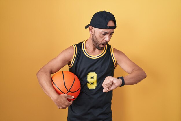 Uomo calvo di mezza età che tiene palla da basket su sfondo giallo guardando il tempo dell'orologio preoccupato, impaurito di arrivare in ritardo