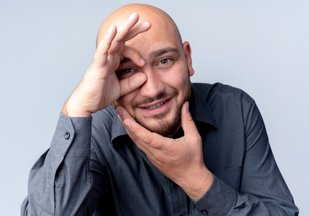 Uomo calvo giovane sorridente della call center che tocca il mento e che fa gesto di sguardo davanti isolato sulla parete bianca