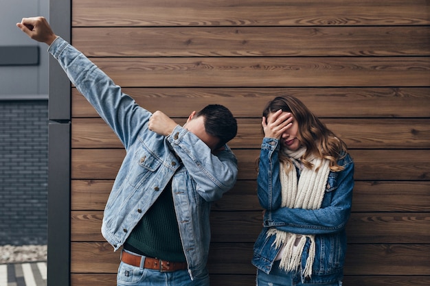 Uomo castana in abbigliamento denim scherzare sulla parete di legno. Ragazza stanca in sciarpa lavorata a maglia lunga che copre gli occhi con la mano.