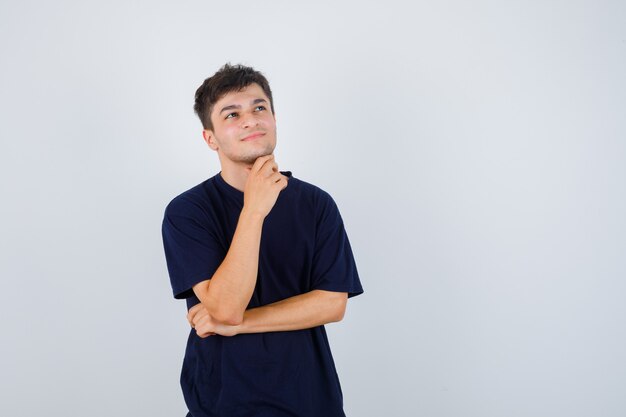 Uomo castana in maglietta che tiene la mano sotto il mento e che sembra pensieroso, vista frontale.