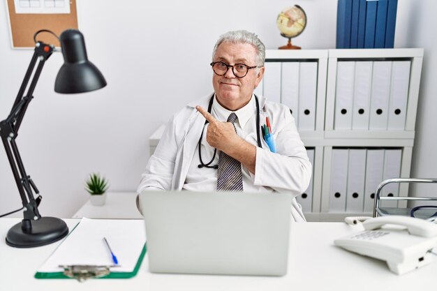 Uomo caucasico anziano che indossa l'uniforme del medico e lo stetoscopio presso la clinica che punta da parte preoccupato e nervoso con l'indice preoccupato e l'espressione sorpresa