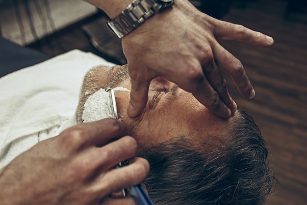 Uomo caucasico barbuto senior bello laterale di vista superiore laterale del primo piano che ottiene governare barba nel parrucchiere moderno.