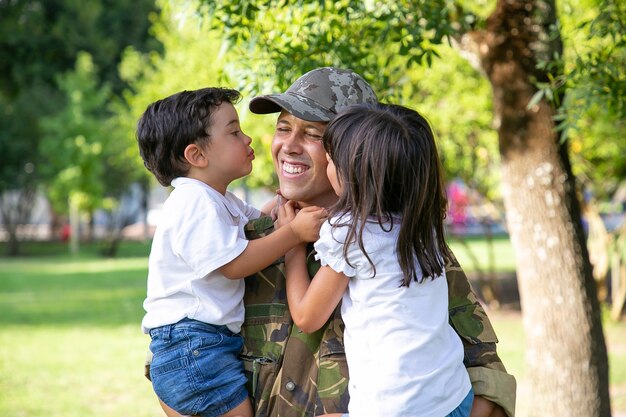 Uomo caucasico che tiene i bambini e sorride. Bambini svegli felici che abbracciano e baciano il padre di mezza età in uniforme militare. Papà che torna dall'esercito. Ricongiungimento familiare, paternità e concetto di ritorno a casa