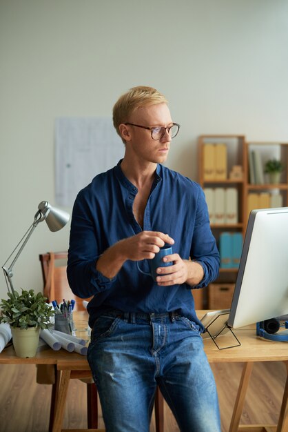 Uomo caucasico creativo che sta davanti allo scrittorio in ufficio, tenendo tazza e distogliere lo sguardo