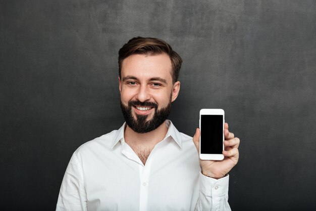 Uomo caucasico del brunette che mostra smartphone sulla dimostrazione della macchina fotografica o che fa pubblicità all'aggeggio sopra lo spazio della copia della grafite