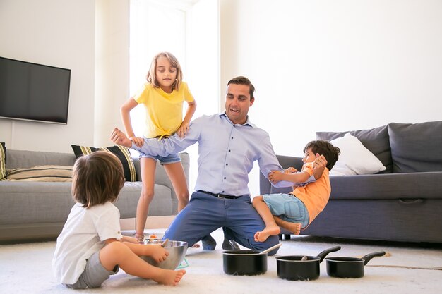 Uomo caucasico felice che gioca con i bambini e che mostra la forza. Bambini allegri che si divertono insieme nel soggiorno sul tappeto. Pentole e ciotola per selvaggina. Infanzia, fine settimana e concetto di attività domestica