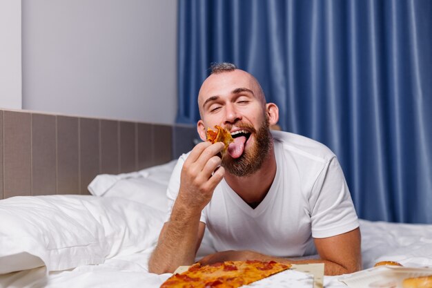 Uomo caucasico felice che mangia fast food a casa in camera da letto sul letto L'uomo ha ordinato cibo da asporto online e mangia pizza e hamburger in camera confortevole