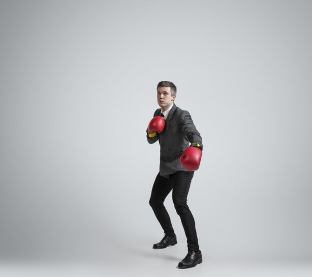 Uomo caucasico in abiti da ufficio boxe con due guanti rossi su sfondo grigio.