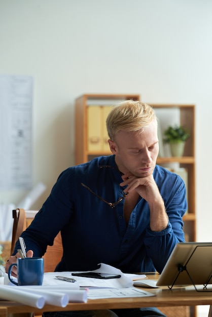 Uomo caucasico in camicia casual seduto alla scrivania in ufficio e guardando tablet