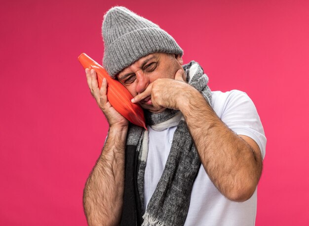 Uomo caucasico malato adulto dispiaciuto con sciarpa intorno al collo che indossa cappello invernale pulisce il naso con la mano e tiene una bottiglia di acqua calda isolata sul muro rosa con spazio di copia