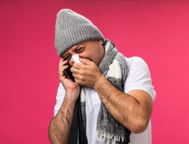 Uomo caucasico malato adulto dispiaciuto con sciarpa intorno al collo che indossa cappello invernale pulisce il naso con un tovagliolo parlando al telefono isolato sul muro rosa con spazio di copia