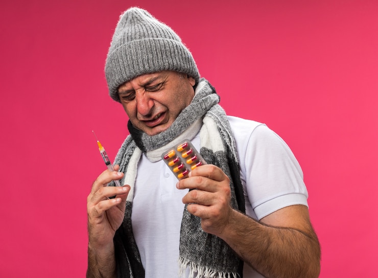 Foto gratuito uomo caucasico malato adulto dolorante con sciarpa intorno al collo che indossa cappello invernale tenendo siringa e blister di medicina isolato sul muro rosa con spazio di copia