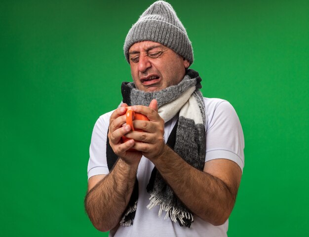 uomo caucasico malato adulto triste con la sciarpa intorno al collo che porta la tazza della tenuta del cappello di inverno isolata sulla parete verde con lo spazio della copia