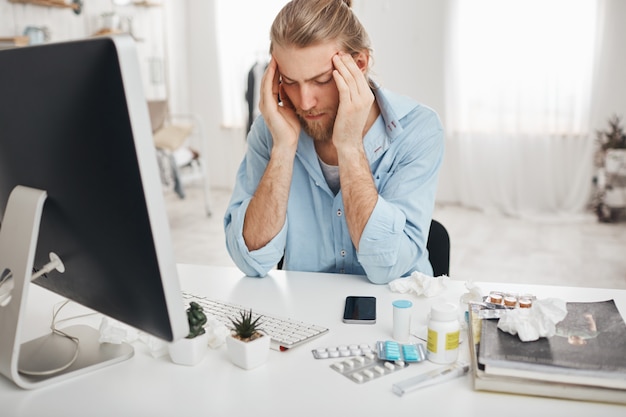 Uomo caucasico malato seduto in ufficio, stringendo le tempie a causa del mal di testa, lavorando sul computer, guardando lo schermo con un'espressione dolorosa sul viso, cercando di concentrarsi, circondato dalla medicina