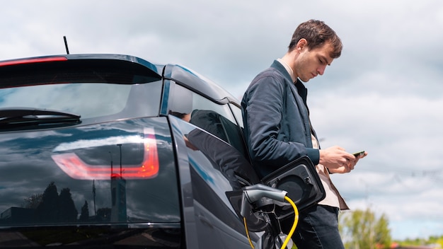 Uomo che carica la sua auto elettrica alla stazione di ricarica e utilizza lo smartphone