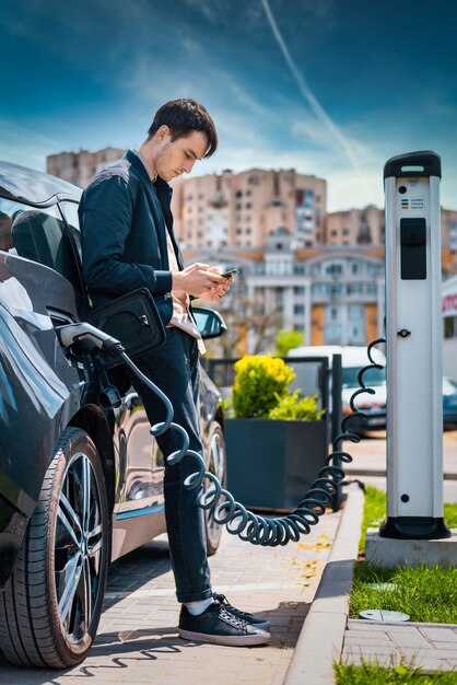 Uomo che carica la sua auto elettrica alla stazione di ricarica e utilizza lo smartphone