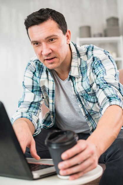 Uomo che digita sul computer portatile e prendendo tazza di bevanda a casa