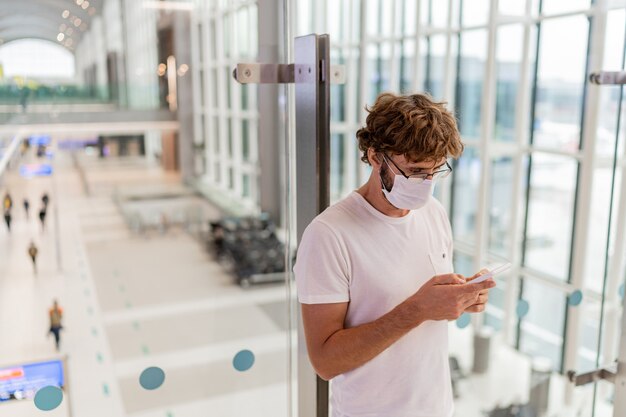 Uomo che indossa la maschera per il viso in aeroporto e utilizza lo smartphone