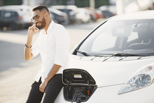 Uomo che parla al cellulare mentre aspetta l'auto elettrica. Stazione di ricarica, auto ecologiche. etnia araba.