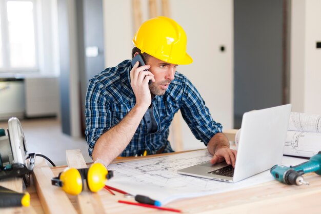 Uomo che parla al telefono cellulare e utilizza il computer portatile sul lato della costruzione