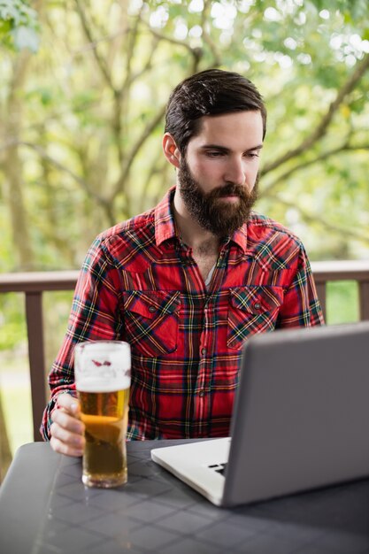 Uomo che per mezzo del computer portatile con bicchiere di birra sulla tavola